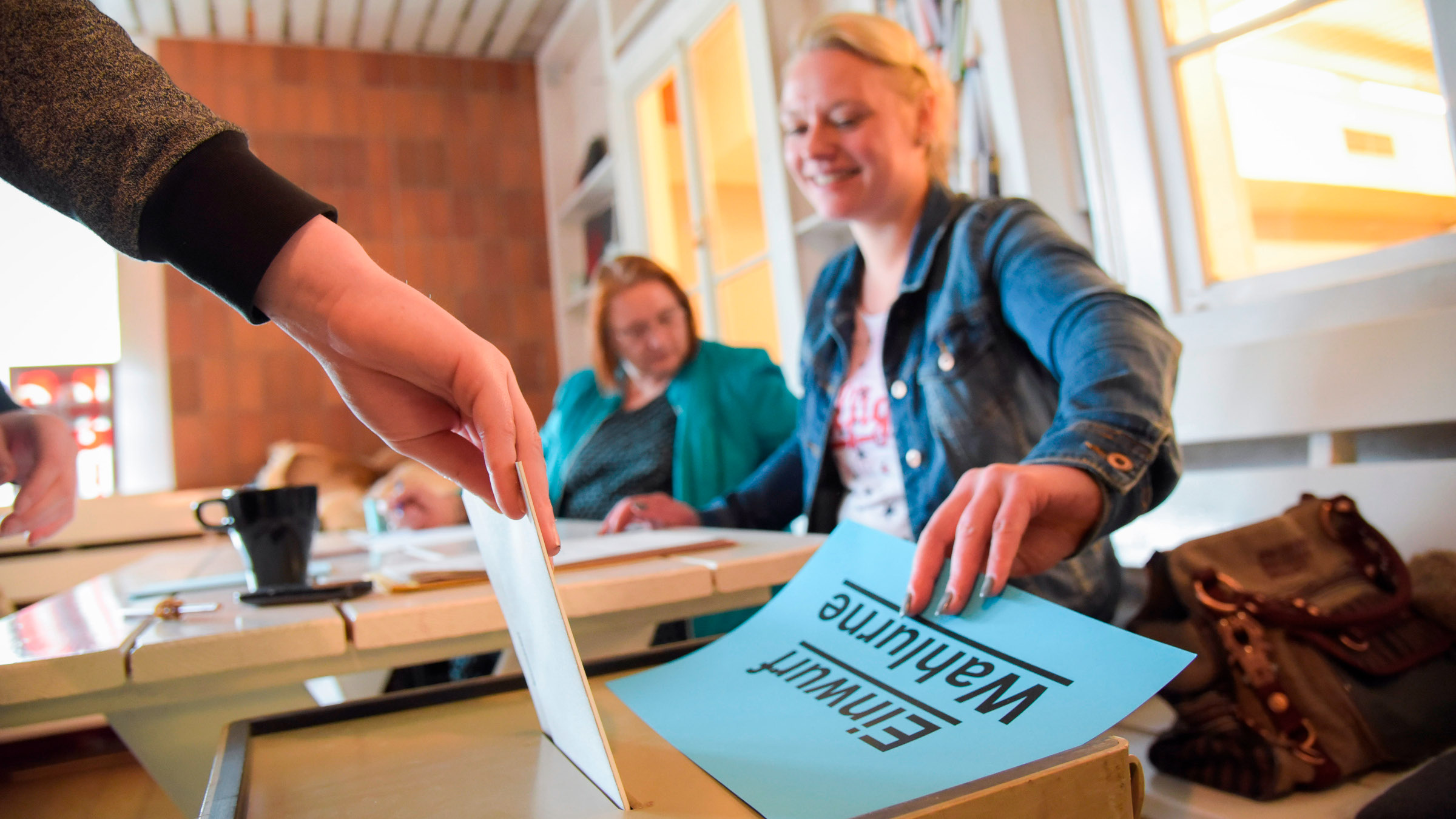 Ein Wähler wirft seinen Stimmzettel in eine Wahlurne. Im Hintergrund sind zwei Wahlhelfer*innen zu sehen.