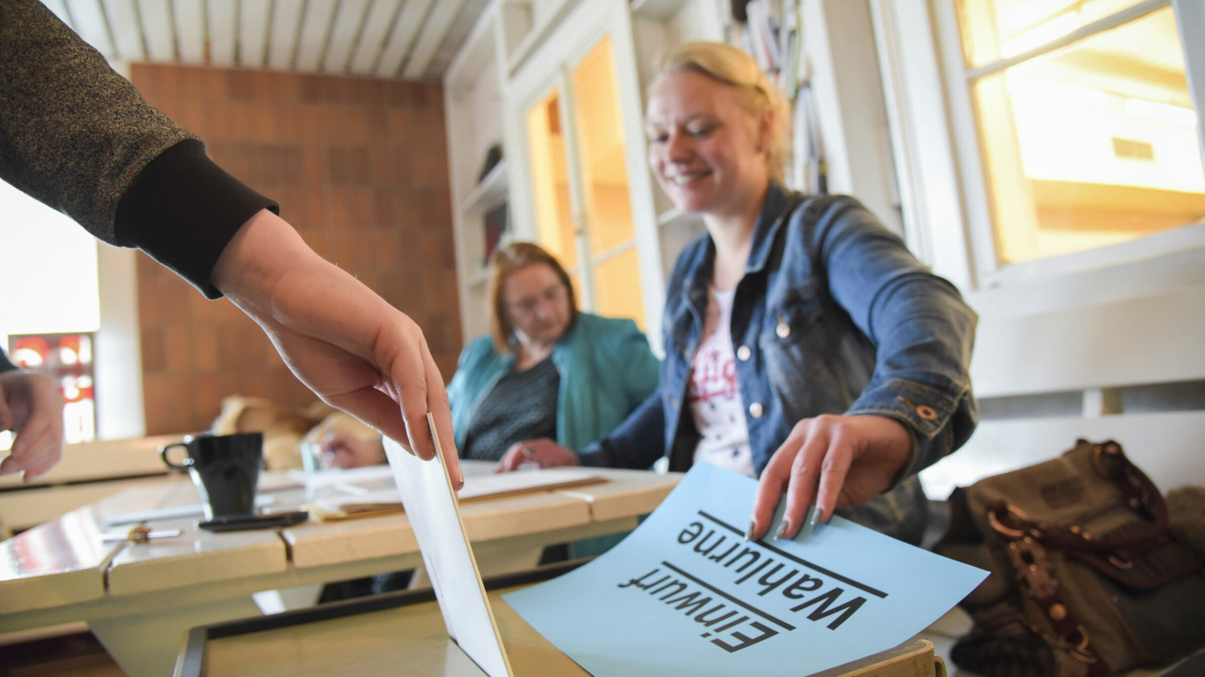 Person gibt einen Stimmzettel in eine Wahlurne, im Hintergrund sitzen zwei Frauen am Tisch und lächeln, während sie den Prozess beobachten.